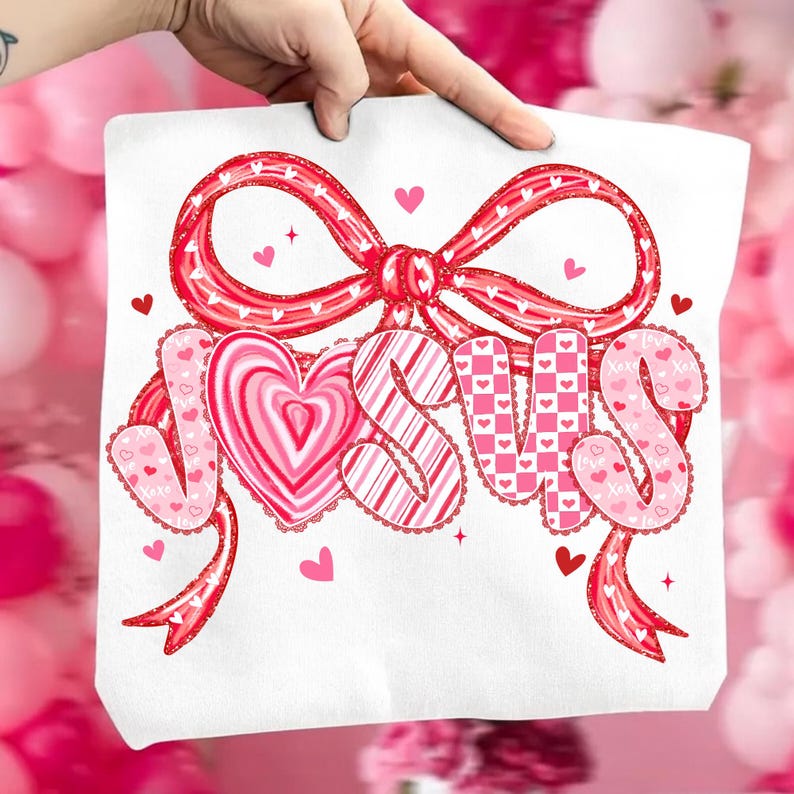 May include: White tote bag featuring the word "JESUS" in pink and red lettering, with a heart-shaped design for the letter "O". The design includes a large red bow and small hearts, perfect for Valentine's Day or religious gifts.