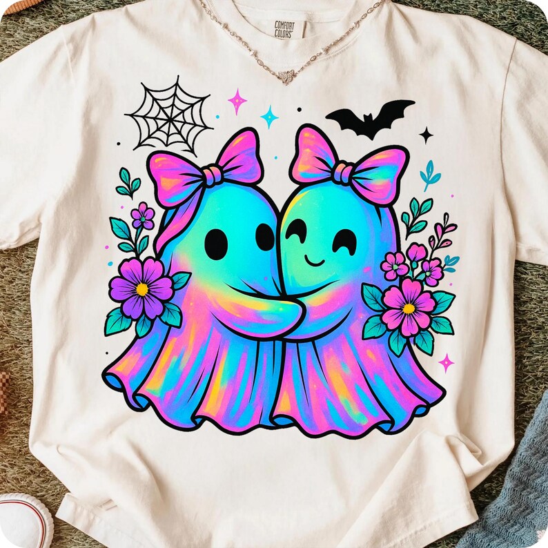 May include: Cream-colored t-shirt featuring a colorful graphic of two hugging ghosts with bows, surrounded by flowers, a spiderweb, and a bat. The shirt has the words "Comfort Colors" printed on the top.