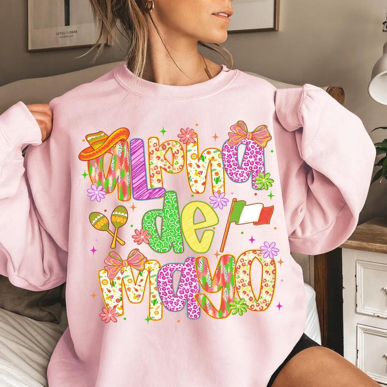 K&ouml;nnte beinhalten: Hellrosa Sweatshirt mit dem Text "Alpha de Mayo" in bunten, gemusterten Buchstaben. Das Design beinhaltet einen Sombrero, Maracas, Schleifen, Blumen und die mexikanische Flagge. Das Sweatshirt ist ein lang&auml;rmeliges Rundhals-Sweatshirt.