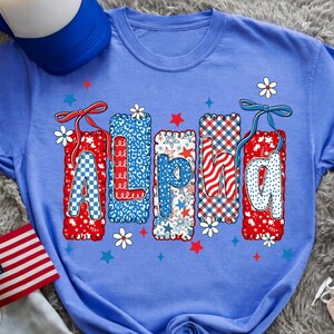 May include: A light blue t-shirt with the word "Alpha" in a patriotic design. The letters are decorated with patterns of red, white, and blue, including stars, checks, and floral accents. A blue and white baseball cap is visible.