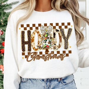 May include: White sweatshirt with a festive "Howdy Christmas" design. The design features a Christmas tree, cow print letters, and a checkered pattern. The text is in brown and gold colors, with a touch of sparkle.
