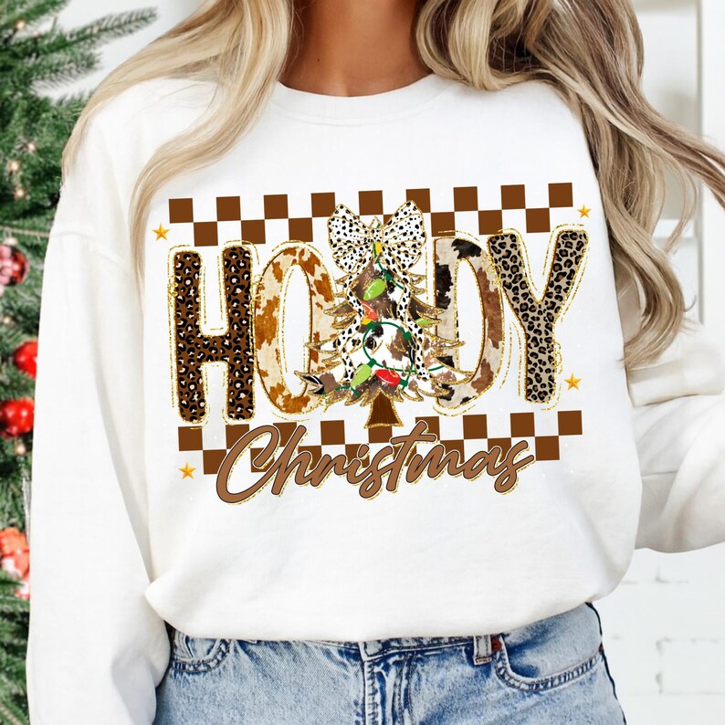 May include: White sweatshirt with a festive "Howdy Christmas" design. The design features a Christmas tree, cow print letters, and a checkered pattern. The text is in brown and gold colors, with a touch of sparkle.