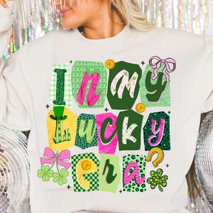May include: Cream-colored sweatshirt with the text "I'm in my lucky era" in a colorful, patchwork design. The design includes green, pink, yellow, and black elements, with shamrocks, bows, and coins.