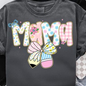 K&ouml;nnte beinhalten: Dunkelgraues Sweatshirt mit dem Wort "Mama" in einem farbenfrohen, floralen und gemusterten Design. Der Buchstabe "M" hat ein Blumenmuster, das "A" hat ein rosa Vichy-Muster, und die anderen Buchstaben haben ein rosa-blaues Blumenmuster. Eine gro&szlig;e Schleife mit Bleistift-Design ist darunter.