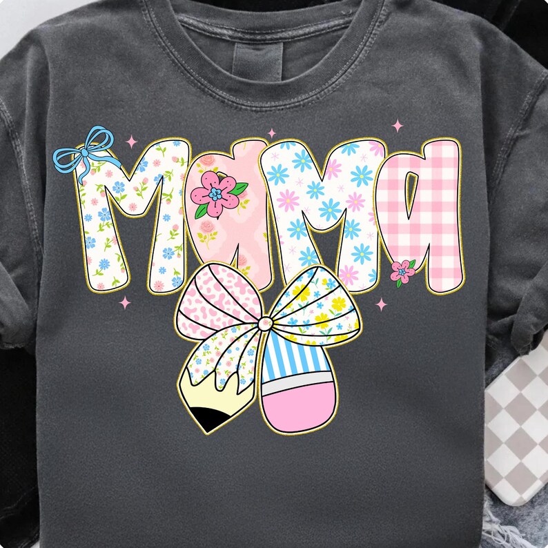 K&ouml;nnte beinhalten: Dunkelgraues Sweatshirt mit dem Wort "Mama" in einem farbenfrohen, floralen und gemusterten Design. Der Buchstabe "M" hat ein Blumenmuster, das "A" hat ein rosa Vichy-Muster, und die anderen Buchstaben haben ein rosa-blaues Blumenmuster. Eine gro&szlig;e Schleife mit Bleistift-Design ist darunter.