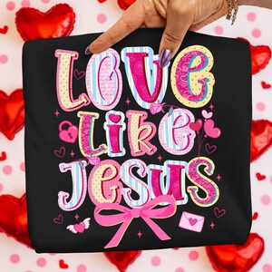 May include: Black t-shirt with the phrase "LOVE LIKE JESUS" in a colorful, whimsical font. The design includes hearts, a lock, an envelope, and a pink bow. The background features red heart-shaped balloons.