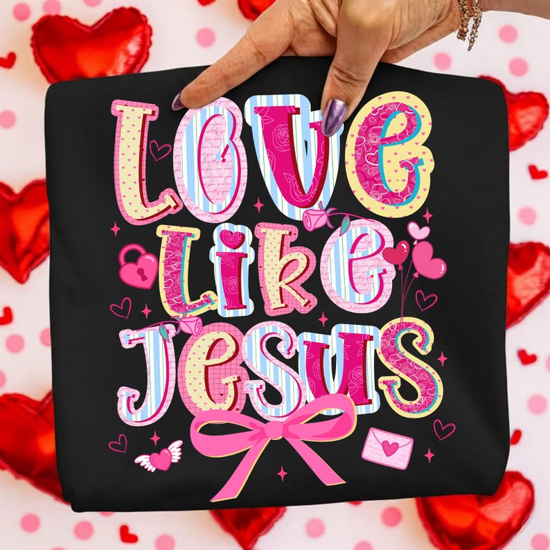 May include: Black t-shirt with the phrase "LOVE LIKE JESUS" in a colorful, whimsical font. The design includes hearts, a lock, an envelope, and a pink bow. The background features red heart-shaped balloons.