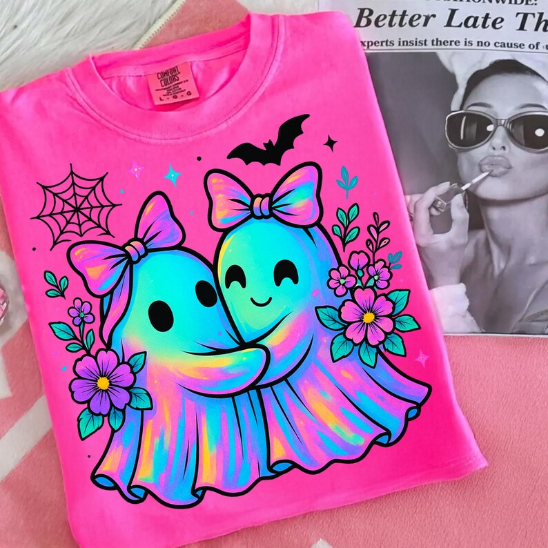 May include: A bright pink t-shirt featuring a graphic of two hugging ghosts with bows, surrounded by flowers, a spiderweb, and a bat. The ghosts are a mix of blue, yellow, and pink. The shirt has a Comfort Colors tag.