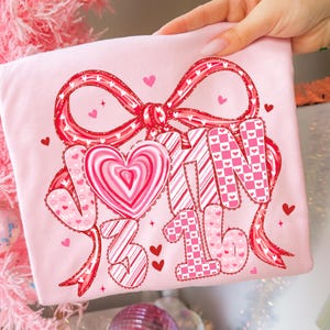 May include: A light pink t-shirt featuring a large red bow with white polka dots and a heart-shaped candy. The shirt has the words "V-Day 3/16" in a mix of patterns and fonts, with small hearts scattered around the design.