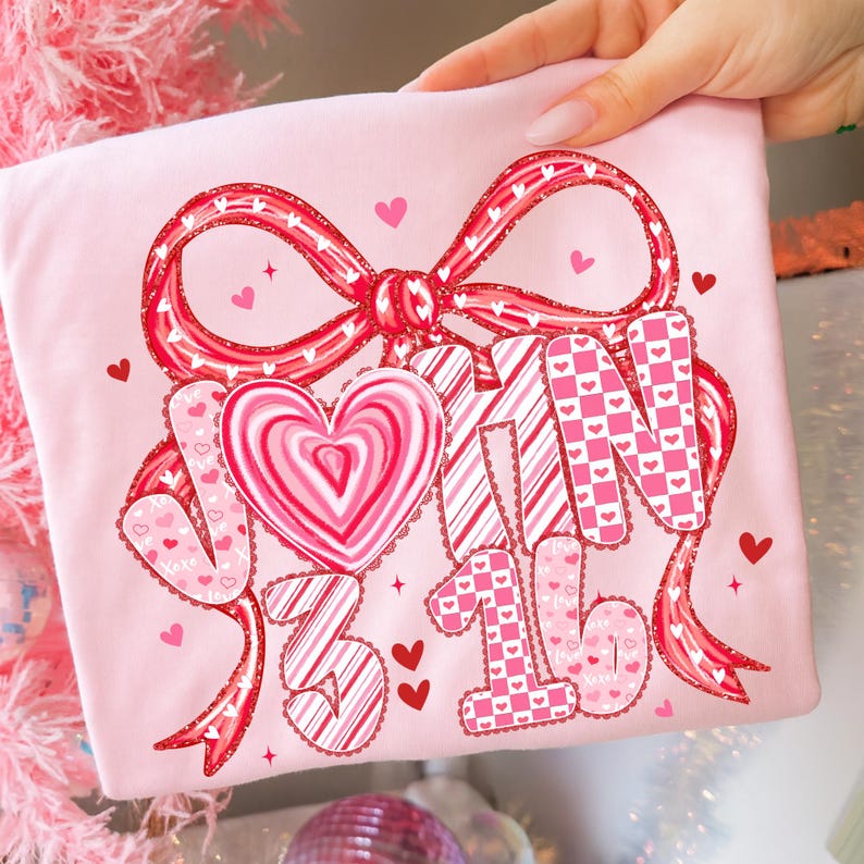 May include: A light pink t-shirt featuring a large red bow with white polka dots and a heart-shaped candy. The shirt has the words "V-Day 3/16" in a mix of patterns and fonts, with small hearts scattered around the design.