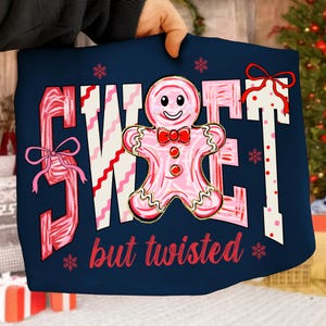May include: Navy blue cushion featuring the words "SWEET" and "but twisted" in red and pink. A pink gingerbread man with a red bow tie is in the center. Pink bows and snowflakes decorate the design.