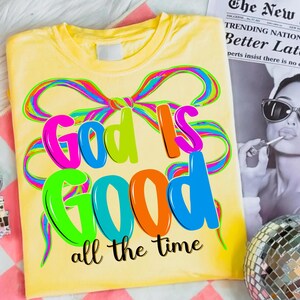May include: Yellow t-shirt with the phrase "God Is Good" in colorful bubble letters and a rainbow ribbon design. The words "all the time" are printed below in black script.