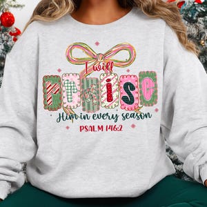 May include: A light gray sweatshirt with the words "I will Praise Him in every season" in a colorful, decorative font. The design includes a bow and floral accents, with the Bible verse Psalm 146:2.