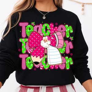 Peut inclure: Sweat-shirt noir avec un motif festif avec le mot "Teacher" en vert et rose, orn&eacute; de flocons de neige et d'un n&oelig;ud du P&egrave;re No&euml;l. Le n&oelig;ud est rose avec des pois blancs et un crayon.