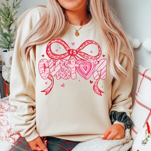 May include: A cream-colored sweatshirt with a large graphic that says "CUSTOM" in pink and white. The word is decorated with hearts and a bow. The sweatshirt is a casual style.