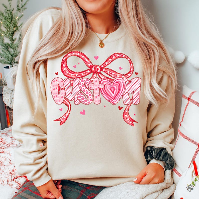 May include: A cream-colored sweatshirt with a large graphic that says "CUSTOM" in pink and white. The word is decorated with hearts and a bow. The sweatshirt is a casual style.