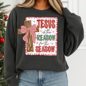 May include: Dark gray sweatshirt featuring a graphic with a cross and the text "JESUS is the REASON for the SEASON" in red, green, and white. The cross has a red and white plaid bow. A festive holiday-themed apparel item.