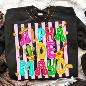 May include: Black sweatshirt with a colorful graphic that reads "Alpha De Mayo." The design includes a sombrero, cactus, guitar, maracas, and a taco. The text is in various bright colors, with pink vertical stripes in the background.