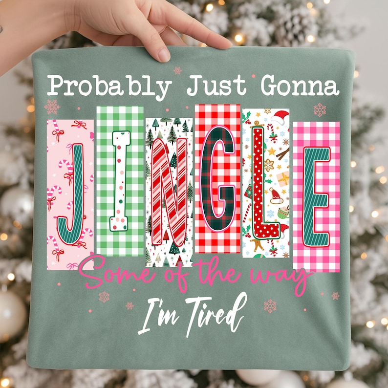 May include: A sage green Christmas-themed t-shirt with the text "Probably Just Gonna Jingle Some of the Way I'm Tired." The word "JINGLE" is spelled out in colorful, patterned blocks with holiday designs.