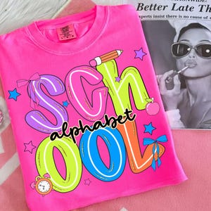 May include: A vibrant pink t-shirt featuring the words "SCHOOL ALPHABET" in bold, multi-coloured letters. The design includes a pencil, stars, an apple, and a clock. The shirt is made of a soft material, ideal for the classroom.