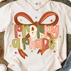 May include: Cream-colored t-shirt with the words "PREPPY ALPHABET" in a colorful, outlined font. The design includes a red and green ribbon bow and small star accents. The shirt is made of Comfort Colors material.