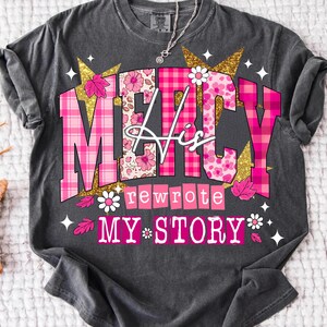 Op de afbeelding: Donkergrijs t-shirt met de tekst "MERCY His rewrote MY STORY" in roze, wit en goud. Het ontwerp bevat bloemen- en ruitpatronen, sterren en bladeren. Een zilveren ketting is zichtbaar.