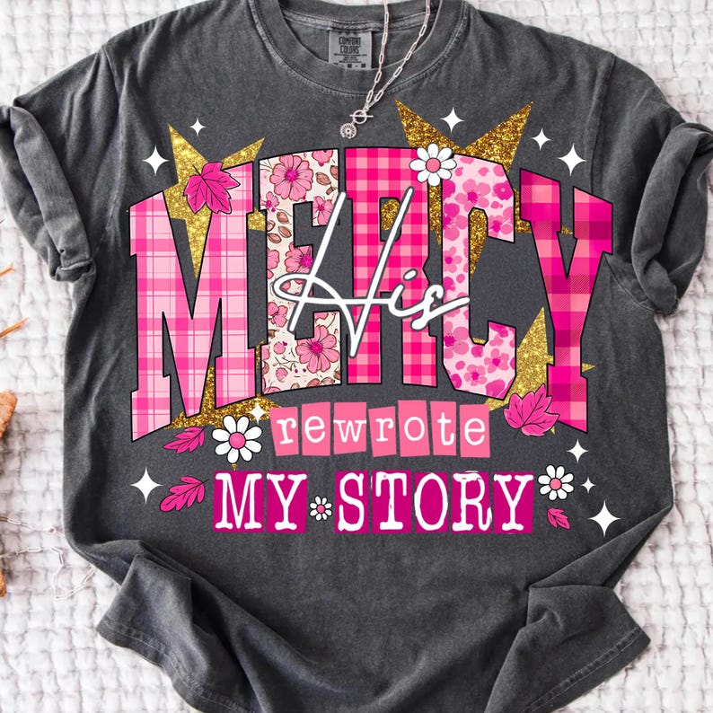Op de afbeelding: Donkergrijs t-shirt met de tekst "MERCY His rewrote MY STORY" in roze, wit en goud. Het ontwerp bevat bloemen- en ruitpatronen, sterren en bladeren. Een zilveren ketting is zichtbaar.