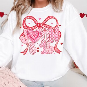 May include: White sweatshirt with a large red bow and the text "John 3:16" in pink. The design includes hearts and patterns, creating a Valentine's Day theme. The sweatshirt is a casual, comfortable style.