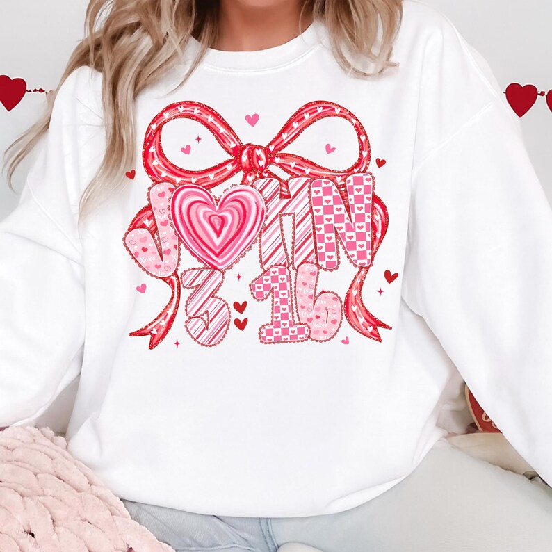 May include: White sweatshirt with a large red bow and the text "John 3:16" in pink. The design includes hearts and patterns, creating a Valentine's Day theme. The sweatshirt is a casual, comfortable style.