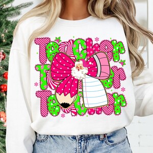 Peut inclure: Sweat-shirt blanc avec un motif graphique color&eacute;. Le motif pr&eacute;sente le mot "Teacher" en rose et vert, un n&oelig;ud rose &agrave; pois blancs, une illustration du P&egrave;re No&euml;l, un crayon et des flocons de neige.