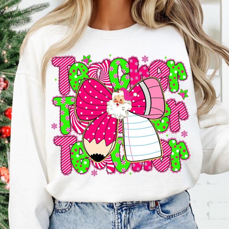 Peut inclure: Sweat-shirt blanc avec un motif graphique color&eacute;. Le motif pr&eacute;sente le mot "Teacher" en rose et vert, un n&oelig;ud rose &agrave; pois blancs, une illustration du P&egrave;re No&euml;l, un crayon et des flocons de neige.