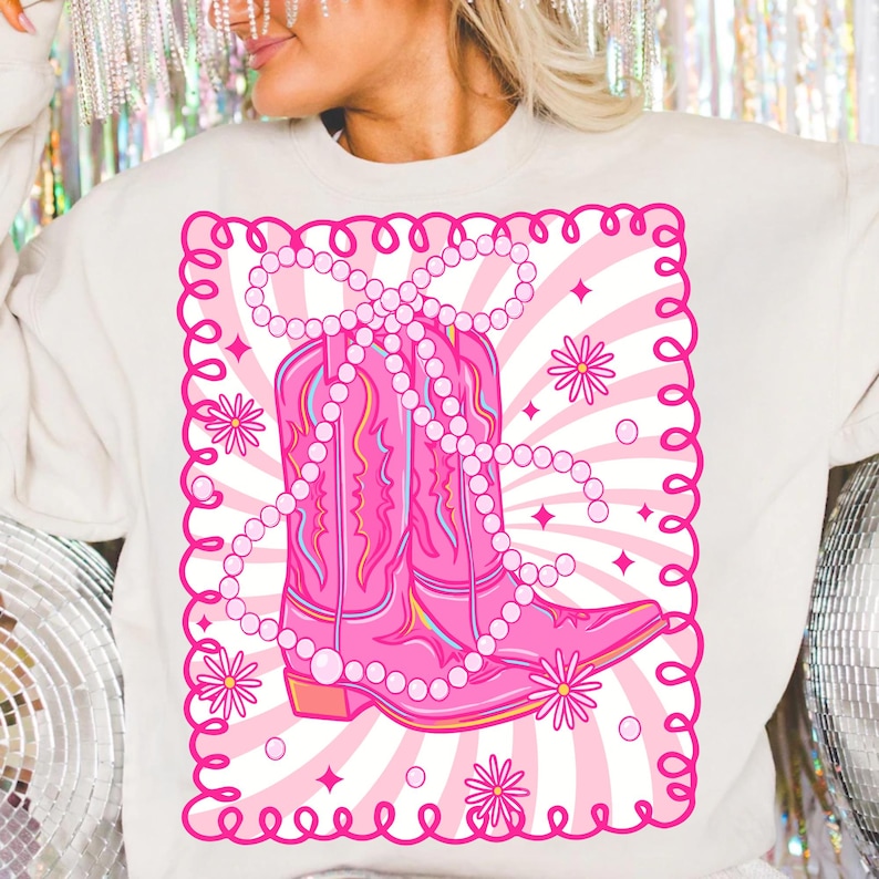 May include: Cream-colored sweatshirt featuring a pink graphic of cowboy boots adorned with a pearl necklace. The boots are set against a pink and white striped background, framed by a pink scalloped border with floral accents.