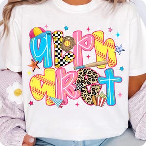 May include: White t-shirt with the words "Girl's About" in colorful, baseball-themed lettering. The design includes a baseball glove, bat, popcorn, and stars. The letters are outlined in various colors.