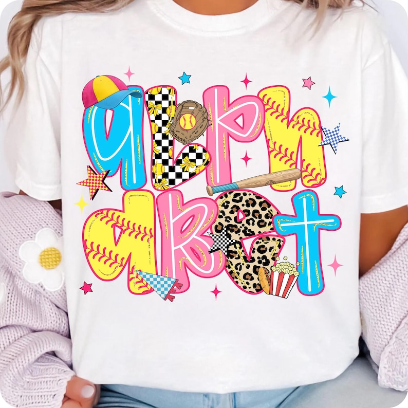 May include: White t-shirt with the words "Girl's About" in colorful, baseball-themed lettering. The design includes a baseball glove, bat, popcorn, and stars. The letters are outlined in various colors.