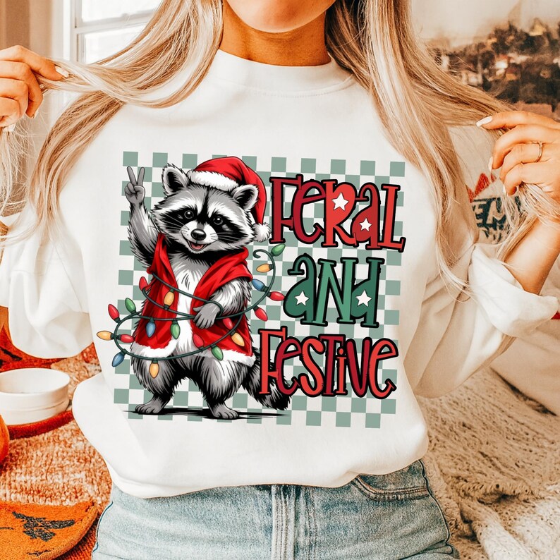 May include: White sweatshirt featuring a raccoon wearing a Santa hat and red vest, holding a string of Christmas lights. The text "FERAL AND FESTIVE" is printed in red and green. The background is a green and white checkered pattern.