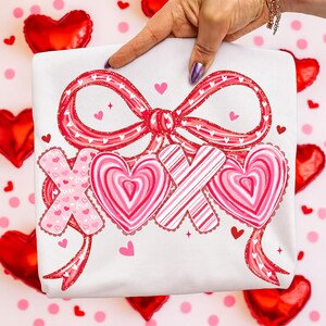 May include: White t-shirt featuring a red bow and the letters XOX, adorned with hearts and stripes. The design incorporates pink hearts and stars, ideal for Valentine's Day or expressing affection. The shirt is held up by a hand.