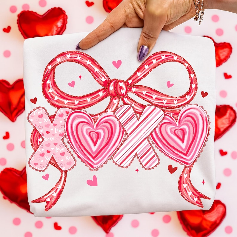 May include: White t-shirt featuring a red bow and the letters XOX, adorned with hearts and stripes. The design incorporates pink hearts and stars, ideal for Valentine's Day or expressing affection. The shirt is held up by a hand.