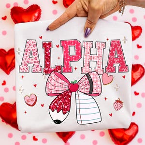 Op de afbeelding: Wit t-shirt met het woord "ALPHA" in roze, met patronen zoals harten en strikken. Een grote strik en een potloodafbeelding staan eronder, samen met harten, aardbeien en de tekst "XOXO".