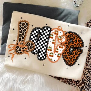May include: A cream-coloured decorative pillow featuring the word "LOVE" in various patterns. The "L" and "E" have a leopard print, the "O" has a checkerboard pattern, and the "V" has a pumpkin and cherry pattern. Black lace trim and orange bows adorn the letters.