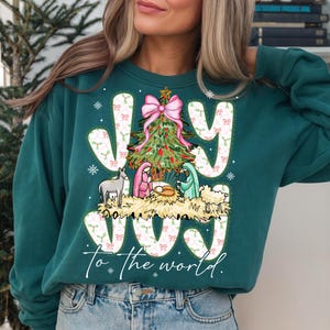 May include: Teal green sweatshirt with a Christmas-themed graphic. The design features the word "JOY" in white, a Christmas tree, and a nativity scene. The phrase "To the world" is written below the graphic.