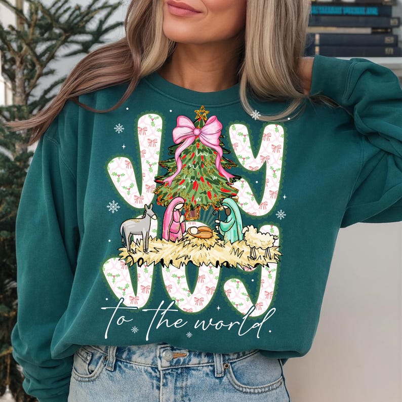 May include: Teal green sweatshirt with a Christmas-themed graphic. The design features the word "JOY" in white, a Christmas tree, and a nativity scene. The phrase "To the world" is written below the graphic.