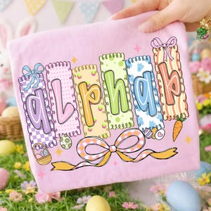 May include: A pink Easter-themed towel with the word "Alpha" in colorful, decorative letters. Each letter is in a different color and design, with Easter eggs, bows, and a carrot. The towel is held by a hand.