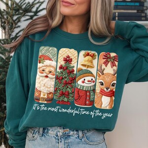 May include: Teal green sweatshirt featuring four Christmas-themed designs: Santa Claus, a Christmas tree, a snowman, and a reindeer. Each design is in a vertical rectangle. The text "It's the most wonderful time of the year" is printed at the bottom.