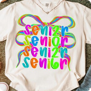 May include: Cream-colored t-shirt with the word "SENIOR" in a colorful, layered design. The word is in a rainbow of colors, with a large, multi-colored bow graphic above. The shirt is a Comfort Colors brand.