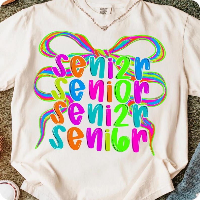May include: Cream-colored t-shirt with the word "SENIOR" in a colorful, layered design. The word is in a rainbow of colors, with a large, multi-colored bow graphic above. The shirt is a Comfort Colors brand.