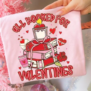 May include: A light pink t-shirt with a Valentine's Day graphic. The graphic features a stack of red books, a candle, hearts, and the text "ALL BOOKED FOR VALENTINES" in red. A red rose and a coffee cup are also included.