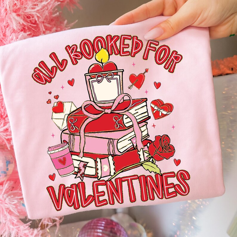 May include: A light pink t-shirt with a Valentine's Day graphic. The graphic features a stack of red books, a candle, hearts, and the text "ALL BOOKED FOR VALENTINES" in red. A red rose and a coffee cup are also included.