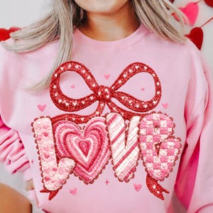 May include: Pink sweatshirt with a red bow and the word "LOVE" embroidered in pink and white. The letters are designed with hearts and stripes. Small heart details are scattered around the design.