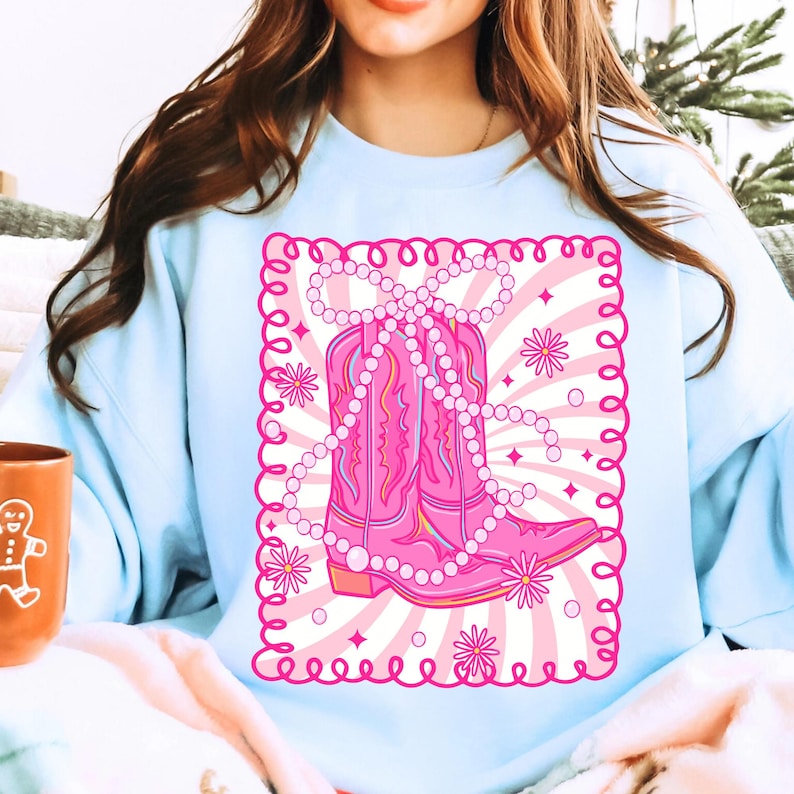 May include: Light blue sweatshirt featuring a pink graphic of cowboy boots adorned with a pearl necklace. The boots are set against a pink and white striped background with floral and star accents, framed by a pink border.