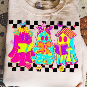May include: A beige sweatshirt featuring a colorful graphic of three ghost figures reading books. The ghosts are in bright neon colors with floral and star accents, set against a black and white checkered border. The design has a playful, whimsical style.